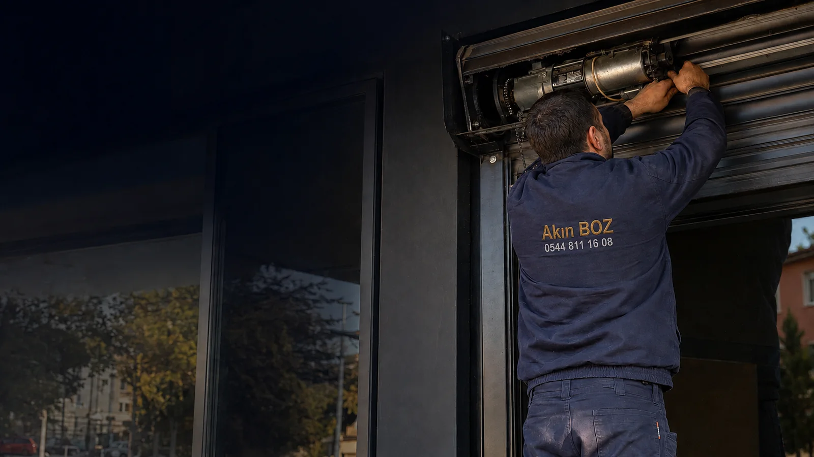 Sırtında Akın Boz ve servis telefonu işlenmiş montla otomatik kepenk motorunun bakımını yapan teknisyen